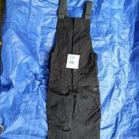 Full front view of black DC bib-style snowboard/ski pants laid flat showing front pockets, adjustable straps, and logo.