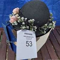 Black straw hat decorated with pink roses and white flowers placed on or near a white mannequin head with a maxsold tag visible.