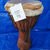 Front view of wooden djembe drum showing natural wood body and animal skin drumhead secured with black rope.