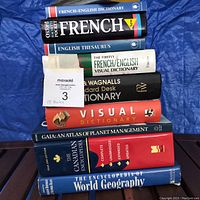 Stack of 10 books including French-English dictionary, English thesaurus, visual dictionaries, standard desk dictionary, atlases, Canadian encyclopedia, and world geography encyclopedia on blue backdrop.