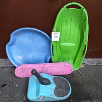 Four winter sleds arranged together: blue saucer, green toboggan, pink floating board, and blue sit-on sled