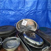 Image showing assortment of large roasting pans, including oval and rectangular pans, some with racks and handles, stack of Pyrex glass bowls with lids, and other cookware items on table with blue backdrop.