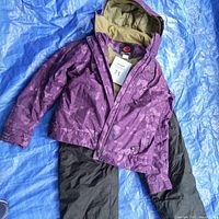 Purple Rossignol ski jacket with hood and beige lining lying on blue tarp with black snow pants underneath.