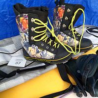 Close-up of pair of Forum limited edition snowboarding boots, men's US size 9, with distinctive colorful print, black leather/synthetic base, and yellow-green laces, alongside parts of the snowboard binding and bag.