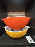 Two large vintage Pyrex mixing bowls stacked; the bottom one yellow-orange with flower pattern and white inside, the top one solid red-orange with white inside and handles