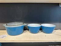 Front view of blue Pyrex casserole dish with lid and two matching blue mixing bowls with white interiors on light wood surface against dark wall.