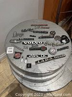 Top view of various original car emblems and badges arranged on a wooden spool showing multiple brand logos and model names.