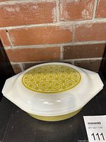 Oval white Pyrex covered casserole dish with yellow geometric basket weave pattern on top of lid against a brick background