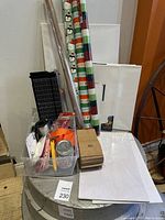 Photo of crafting supplies including wrapping paper rolls, plastic container with scissors, tape and wax kit, small brown paper boxes, and blank canvases grouped together.