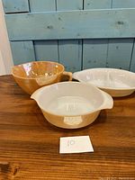 Three Fire King lusterware pieces displayed together on a wood surface against a blue background. Includes a large mixing bowl, a smaller round casserole, and a divided serving dish.