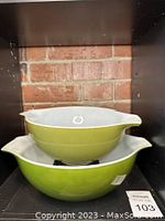 Two green Pyrex Cinderella bowls stacked on each other against brick background, showing size difference and characteristic spouted handles.