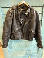 Front view of the men's black leather riding jacket hanging on a wooden hanger in front of a blue wooden backdrop. Shows collar, zipper and pocket details.