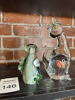 Two Murano glass cat figurines, one clear and one green/brown speckled, displayed against brick background.