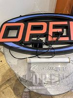 Oval blue and black illuminated open sign with orange text and cords on round table, clear hanging sign below it.