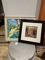 Two framed artworks displayed together on a small round table, one colorful original painting and one sepia-toned print.