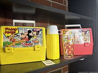 Yellow and red Mickey Mouse lunch boxes on shelf along with yellow thermos and Mickey Mouse glass