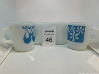 Front view of all four mugs showing two with blue designs (teardrop and flower) and two plain white mugs.