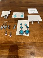 Several pairs of various drop-style earrings displayed on a wooden surface, showing different colors, shapes, and designs including turquoise stones, silver tones, pink accents, and geometric patterns.
