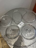 Photo showing five clear glass lids of varied sizes and shapes on a round table, three with knob handles, two flat and unbranded.