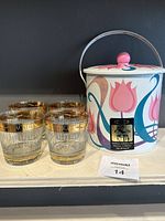 Image of floral decorated vintage ice bucket with metal handle and matching pink lid knob, alongside four short Scotch glasses with gold and black decorative banding on the rims and bases.