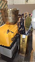 Full view of the brass umbrella stand filled with umbrellas, brass planter bowl on top of yellow equipment, metal boxes below