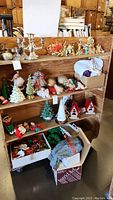Three shelves of assorted holiday decorations including ceramic Christmas trees, Santa, elves, birdhouses, and boxes of additional ornaments and materials.