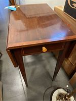 Front angle view of dropleaf table, showing drawer with round knob and scratches on wood surface.