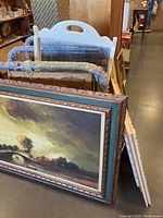 Several framed paintings and mirrors stacked, showing large and smaller framed artworks with ornate and carved wooden frames and two mirrors.