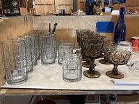 Several clear tall drinking glasses with thick bases and a row of mocha brown goblets and dessert bowls in front.