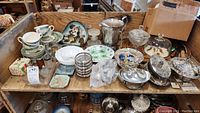 Overview of kitchen decor and dishes on wood shelf, includes teacups, plates, silverplate items, glassware, trinket boxes.