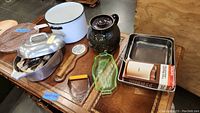 Photo of kitchen items including enamel pot, roasting pan with lid, ceramic teapot, glass dish, and small utensils displayed on a table.