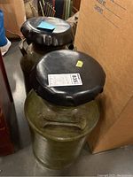 Two milk can stools side by side showing their cushioned black tops and greenish vintage metal cans with handles