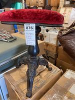 Photo showing full view of antique piano stool with red velvet cushion and ornate metal carved legs, wood and metal construction, has label attached.