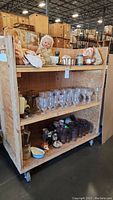 Wooden shelving unit containing depression glass stemware, mugs, assorted small glass and ceramic items, metal molds, and piggy bank shaped like a pig. Some utensils included on shelves.