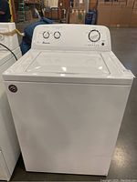 Front view of a white Amana top load washing machine in warehouse, showing washer lid and control panel at rear.