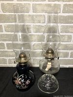 Two oil lamps side by side showing one with black floral base and clear glass base lamp.