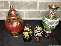 Overall set showing two painted brass decorative eggs with bases, a red lidded brass jar and a colorful dragon decorated brass vase.