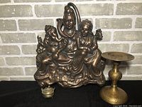Brass plaque depicting Hindu deities Shiva, Parvati, and Ganesha seated, with a small ornate brass candle stand beside it.