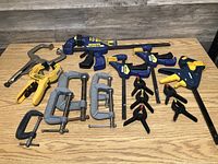 Full lot of assorted clamps including blue and yellow Irwin Quick-Grip clamps, metal C-clamps, spring clamps, and other hand clamps on wooden surface.