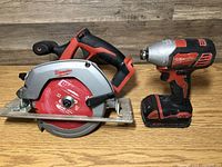 Milwaukee cordless circular saw and cordless impact drill placed on wooden table, showing side profile of saw blade and drill's battery base.