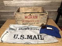 Photo showing wooden crate, U.S. Mail bag, and blue money bag together on a table with wood background.
