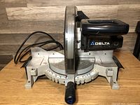 Front left angled view of mitre saw showing blade cover, power cord, and angle adjustment handle.