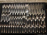 Full layout of the complete silver plated cutlery set showing knives, forks, spoons, and serving pieces arranged on black cloth, visible tarnish and wear.