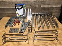 Wide angle photo showing the entire lot of assorted metal hanging hardware on a wooden surface including boxed hooks, metal brackets, clamps, and various hooks