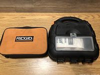 Two carrying cases on wooden surface: one small orange and black Rigid branded case and one black Black & Decker molded plastic case with clear lid and manuals visible inside.