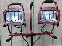 Front view of dual halogen work lights with protective metal cages on adjustable stand, showing foam-padded handles and power cord.