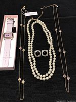 Full view of Kate Spade watch, two heart pendant necklaces, pearl necklace, and Oscar clip-on earrings arranged on black fabric.