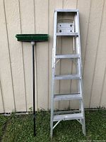 Green push broom and 6 foot aluminum ladder leaning against beige wall on grass.