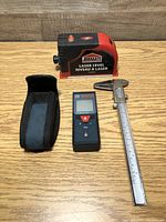 Three measuring devices on a wooden surface: Job Mate laser level, Bosch GLM 40 laser distance meter with carrying pouch, and metal caliper gauge.