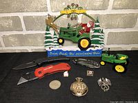 John Deere themed decorative piece with Santa figurine on tractor, two pine trees, and snow base with text 'Santa Parks His John Deere Here', surrounded by various tools and collectible items on table.